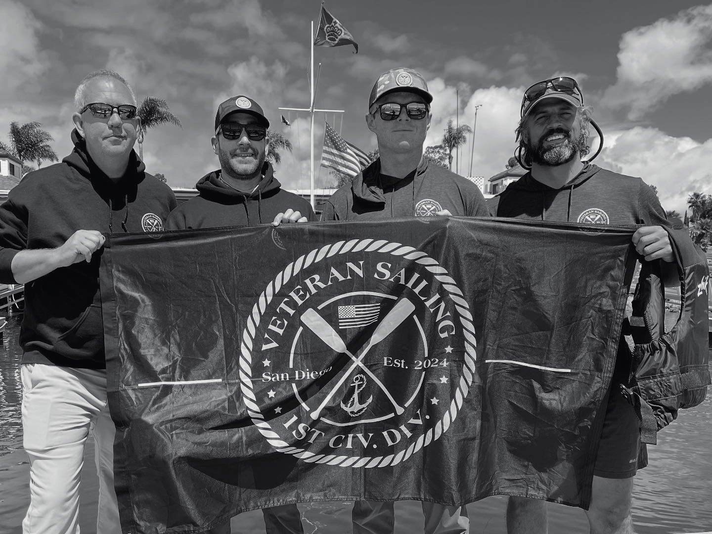 Four men holding a 'Veteran Sailing' flag with a boat in the background.